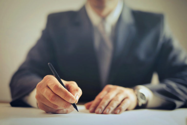 A guy in a suit sitting down holding a pen like he is signing something